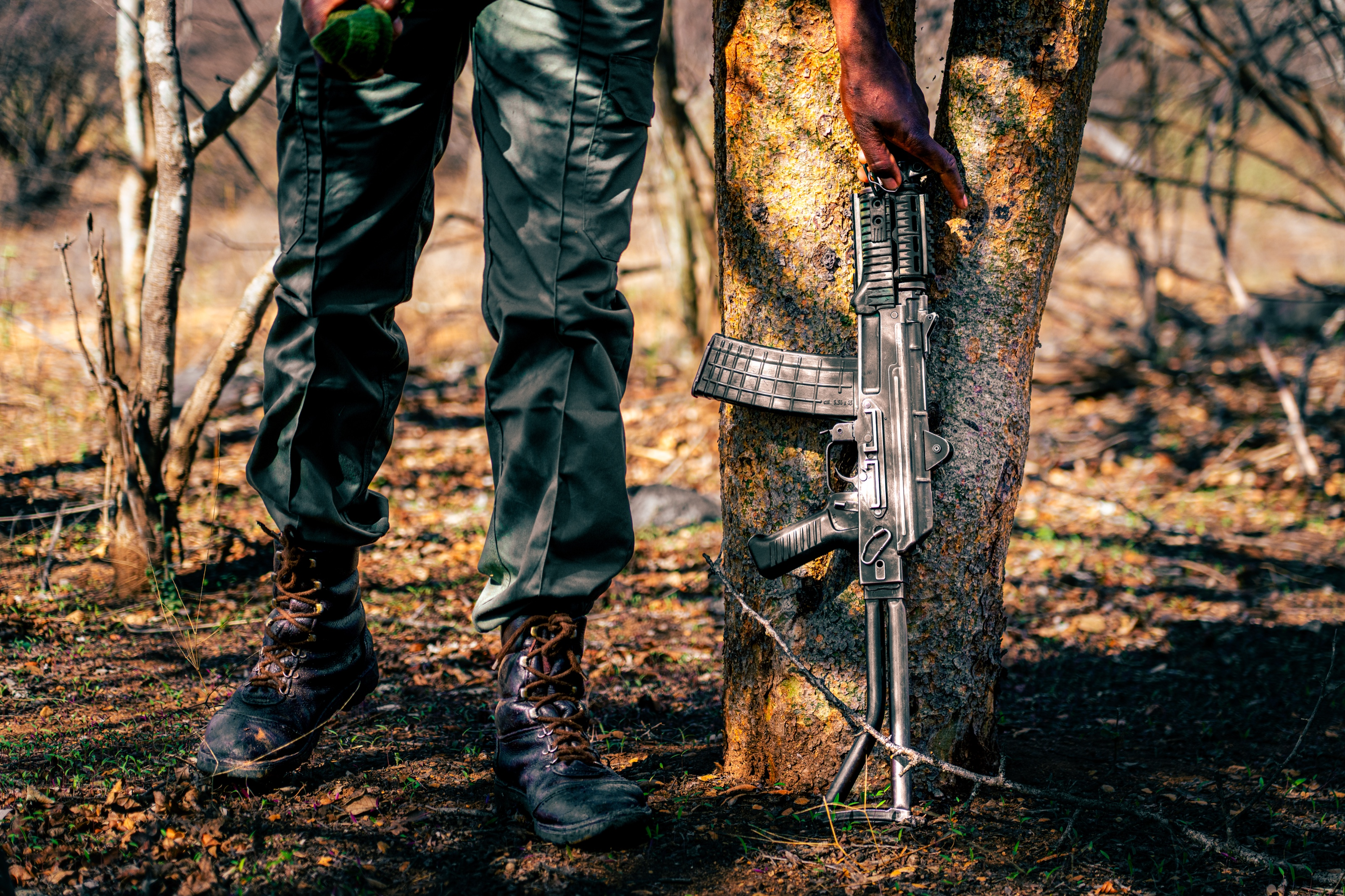 The legs and boots of two people in front of a burnt tree; a rifle rests against the trunk.