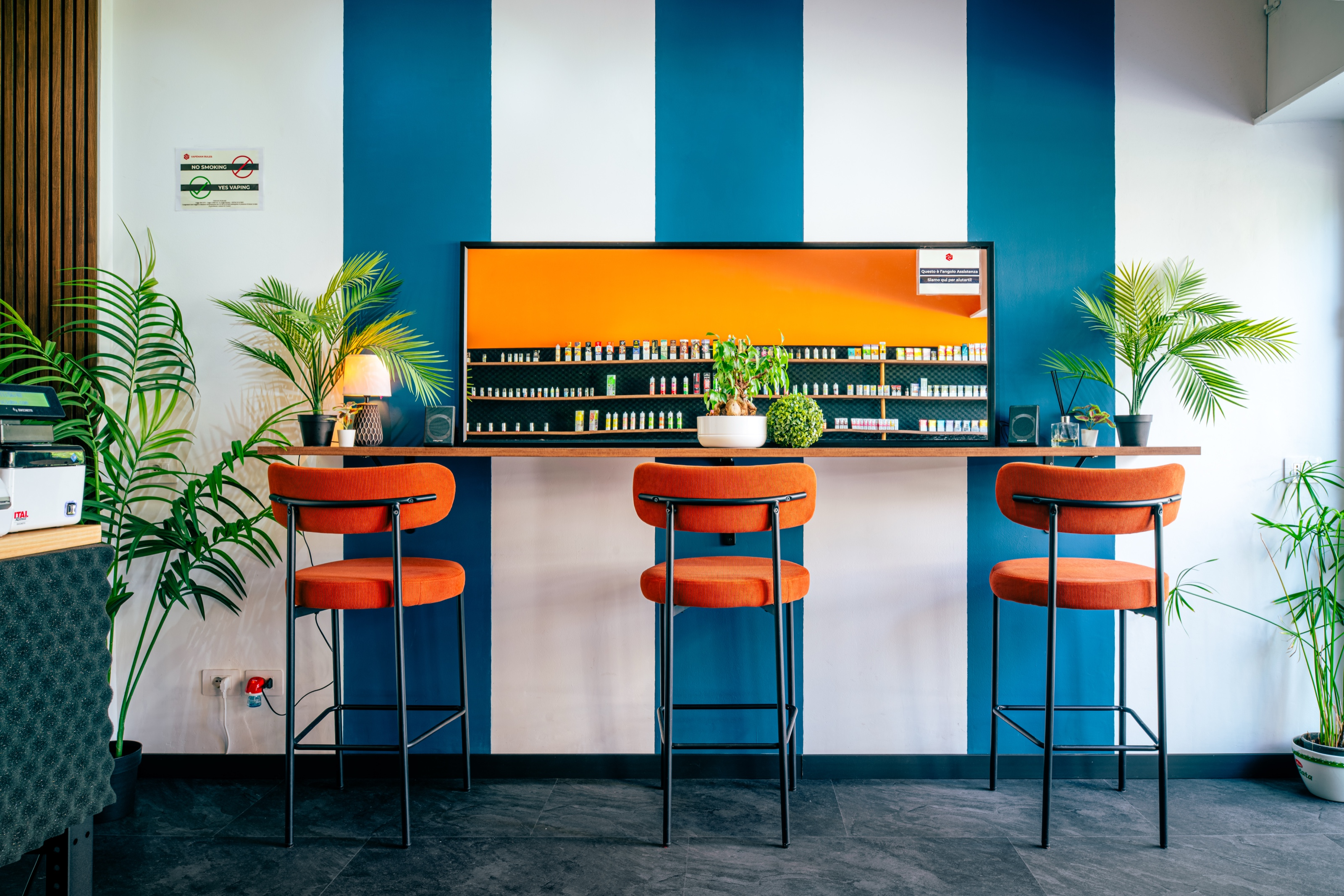 The inside of the new shop: a wall of vertical white and blue stripes, three orange stools at a wooden counter, an orange-lit display case with bottles in a row, potted palms on either side.