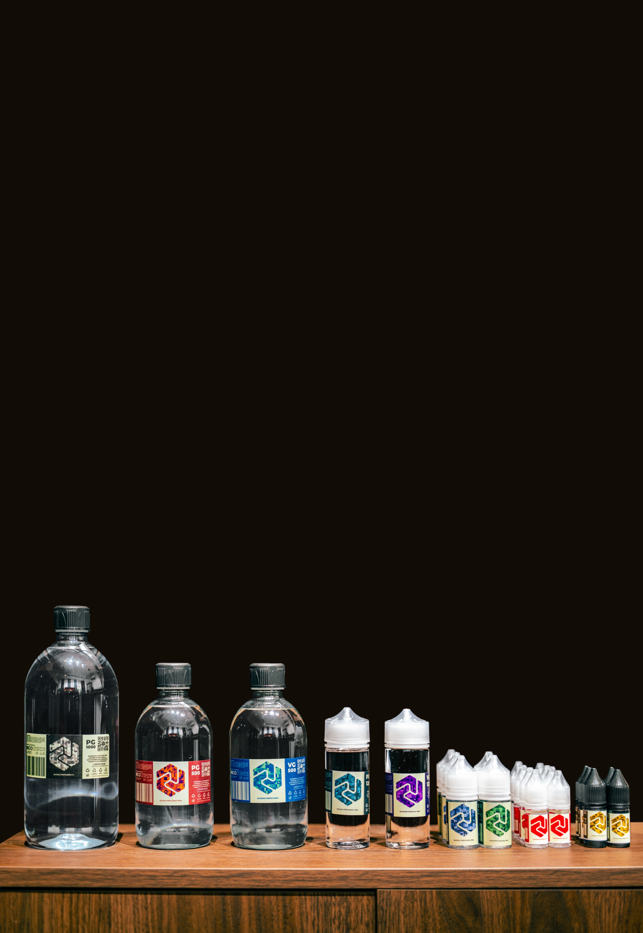 Clear bottles in various sizes lined up on a wooden shelf against a dark background; each carries a label in a different color — green, red, blue, purple, yellow, black — with the Vapeman hexagon.