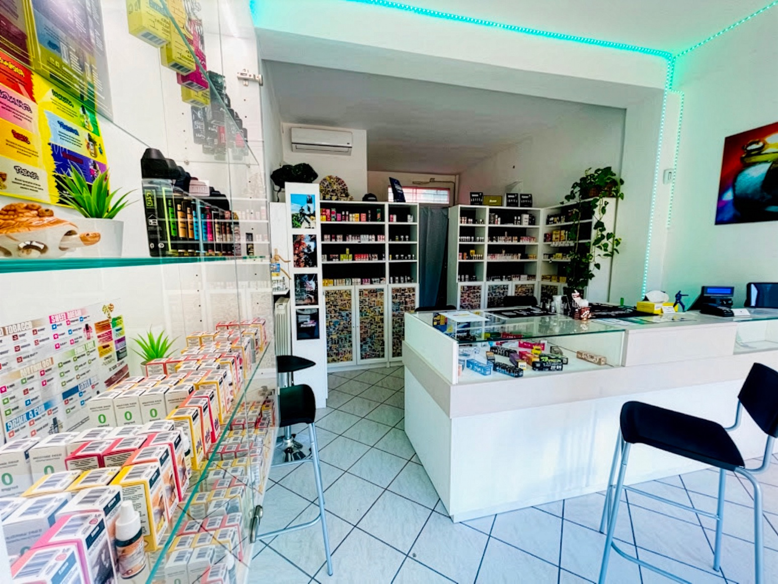 The inside of the old shop: white shelves crammed with bottles and boxes, a display counter, a strip of turquoise LEDs along the ceiling, two black stools on a pale tiled floor.